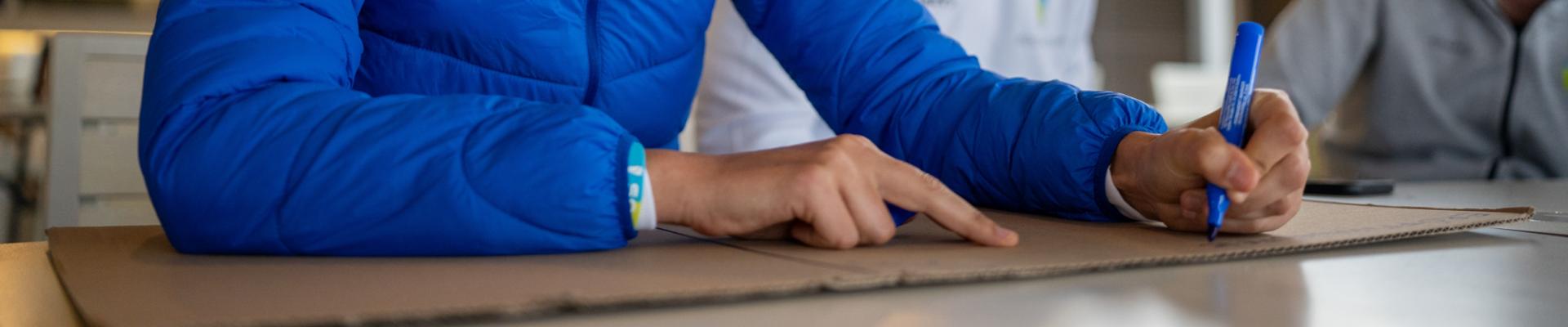 Person in blauer Jacke schreibt mit einem blauen Marker auf einen Karton, w&auml;hrend eine andere Person daneben sitzt.