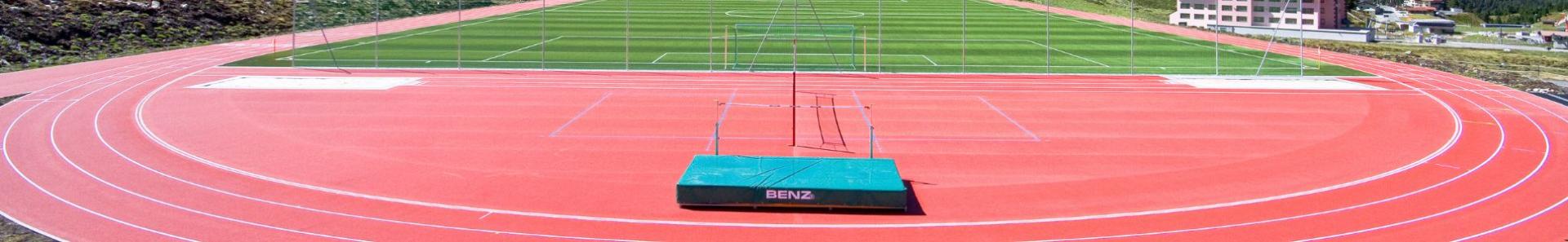 Eine rote Laufbahn umgibt ein gr&uuml;nes Fu&szlig;ballfeld. In der Mitte des Vordergrunds befindet sich eine Hochsprungmatte, wei&szlig;e Linien markieren Bahnen und Felder. Im Hintergrund sind Geb&auml;ude und H&uuml;gel zu sehen.