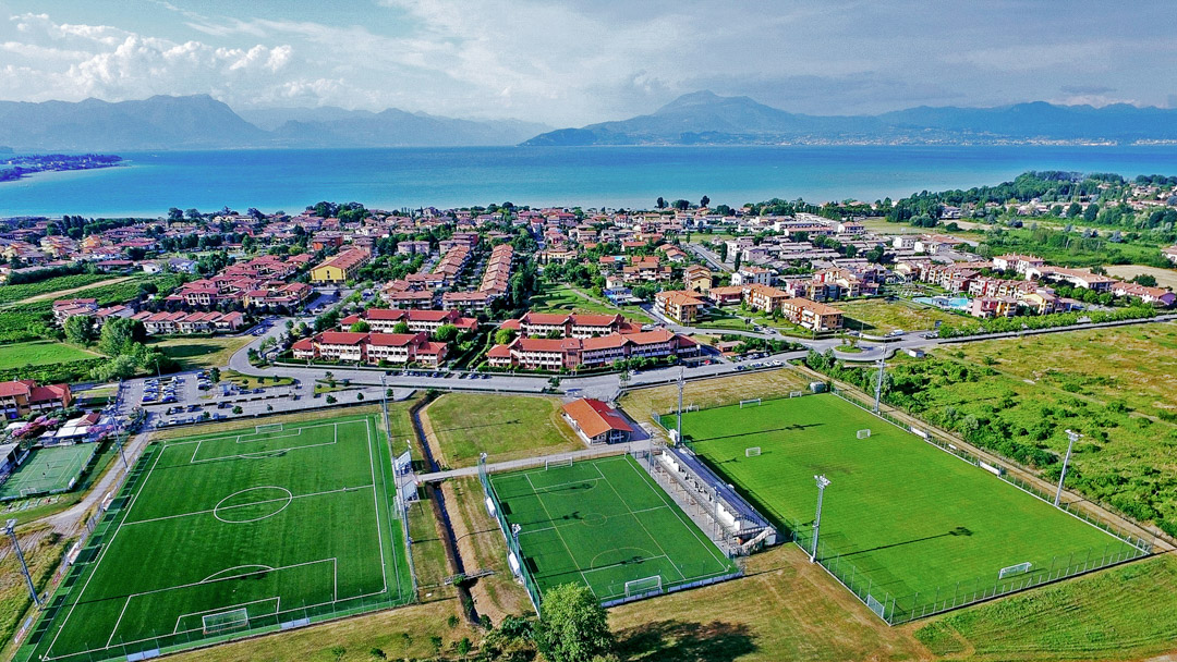 Fußballplätze inmitten einer mediterranen Stadtlandschaft mit Blick auf einen blauen See und umliegende Berge.
